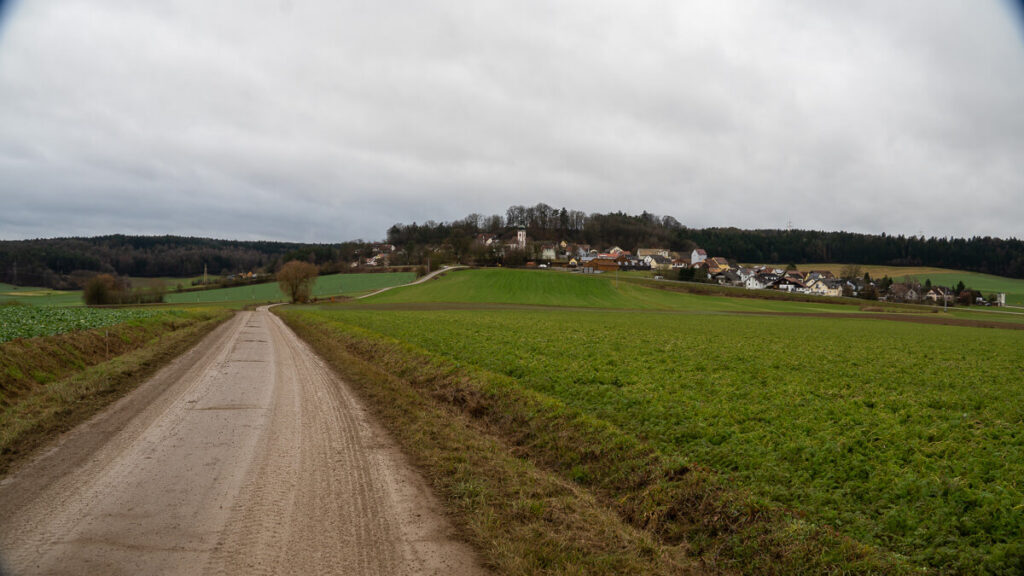 Feldweg durch offene Winterlandschaft, der auf einen Ort am Horizont zuläuft, auf dem Oberpfälzer Jakobsweg